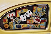 A car window filled with colorful stickers representing different U.S. states and Canada. The stickers display state names, maps, and iconic symbols, such as a maple leaf for Canada and a cartoon moose. The stickers are arranged haphazardly across the window, creating a collage effect.