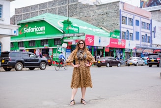 A woman wearing a leopard-print dress poses confidently in the middle of a street. She is surrounded by various vehicles, including cars and a bicycle. In the background, there are shops with green signage and advertisements, one prominently displaying the Safaricom brand.