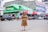 A woman wearing a leopard-print dress poses confidently in the middle of a street. She is surrounded by various vehicles, including cars and a bicycle. In the background, there are shops with green signage and advertisements, one prominently displaying the Safaricom brand.
