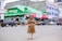 A woman wearing a leopard-print dress poses confidently in the middle of a street. She is surrounded by various vehicles, including cars and a bicycle. In the background, there are shops with green signage and advertisements, one prominently displaying the Safaricom brand.