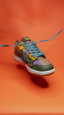 A colorful sneaker with a dynamic design, incorporating shades of green, yellow, purple, and blue. The shoe is positioned against a vivid orange background, with the blue laces prominently extending outward, creating a sense of motion.