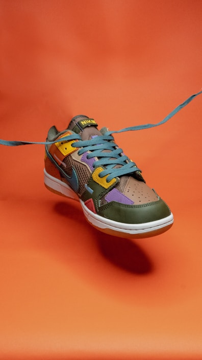 A colorful sneaker with a dynamic design, incorporating shades of green, yellow, purple, and blue. The shoe is positioned against a vivid orange background, with the blue laces prominently extending outward, creating a sense of motion.