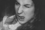 A person making a funny face by scrunching up their eyes and sticking out their tongue. The expression suggests playfulness or defiance. The image is in black and white, which gives it a classic, timeless feel. The person's hair is flowing, and they are wearing a button-up shirt.
