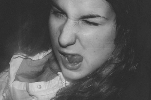 A person making a funny face by scrunching up their eyes and sticking out their tongue. The expression suggests playfulness or defiance. The image is in black and white, which gives it a classic, timeless feel. The person's hair is flowing, and they are wearing a button-up shirt.