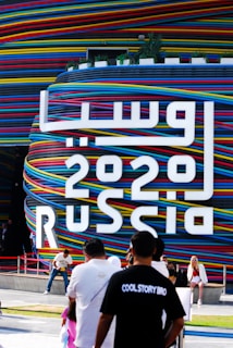 A large, colorful structure is wrapped in horizontal stripes of red, blue, yellow, and black. Bold white text displays the year '2020' alongside what appears to be the word 'Russia' in both Latin and Arabic scripts. Several people are present in the foreground, including a person wearing a black shirt with white text that says 'COOL STORY BRO' and another person in a white top and light pants sitting by the structure.
