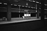 A monochrome image of a deserted train platform at Berlin Hauptbahnhof with an illuminated sign displaying platform numbers 1-2. The environment is dimly lit, featuring industrial design elements like metal pillars and overhead lighting. An information board is centrally located, and there is a trash bin on the left side.