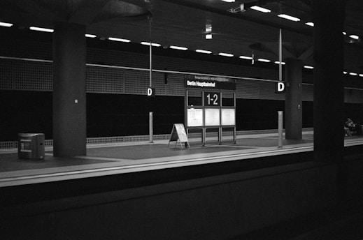 A monochrome image of a deserted train platform at Berlin Hauptbahnhof with an illuminated sign displaying platform numbers 1-2. The environment is dimly lit, featuring industrial design elements like metal pillars and overhead lighting. An information board is centrally located, and there is a trash bin on the left side.