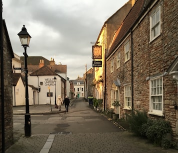 A quaint street scene featuring stone buildings with traditional architecture. The evening sunlight casts a warm glow on the buildings. Two people are walking down the street, and signage indicates a Greek Taverna and a church. The street is quiet and lined with a few potted plants.