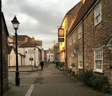 A quaint street scene featuring stone buildings with traditional architecture. The evening sunlight casts a warm glow on the buildings. Two people are walking down the street, and signage indicates a Greek Taverna and a church. The street is quiet and lined with a few potted plants.