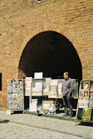 A street art vendor stands in front of a large brick wall with an archway, displaying various framed paintings and colorful artwork. The vendor, wearing a checkered shirt, is positioned next to the artwork, which includes landscapes and abstract designs.