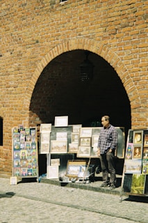 A street art vendor stands in front of a large brick wall with an archway, displaying various framed paintings and colorful artwork. The vendor, wearing a checkered shirt, is positioned next to the artwork, which includes landscapes and abstract designs.