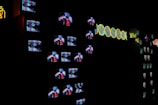 A dark room filled with numerous small television screens displaying colorful images and static. Neon lights form abstract designs and patterns alongside the screens, adding a bright contrast to the darkness.