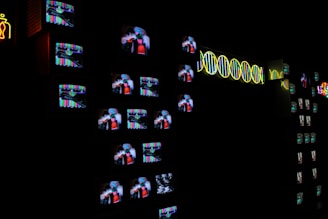 A dark room filled with numerous small television screens displaying colorful images and static. Neon lights form abstract designs and patterns alongside the screens, adding a bright contrast to the darkness.