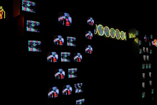A dark room filled with numerous small television screens displaying colorful images and static. Neon lights form abstract designs and patterns alongside the screens, adding a bright contrast to the darkness.