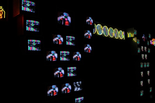 A dark room filled with numerous small television screens displaying colorful images and static. Neon lights form abstract designs and patterns alongside the screens, adding a bright contrast to the darkness.