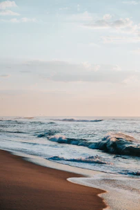 Horizontal photo showing gentle waves on a sandy beach under a pastel sky.
