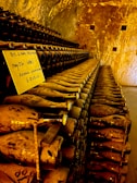A dimly lit cellar filled with rows of dusty wine bottles, all stacked horizontally on shelves. A handwritten label is placed among the bottles, indicating some handwritten notes. The golden-brown tones of the walls and lighting create a warm, aged atmosphere.