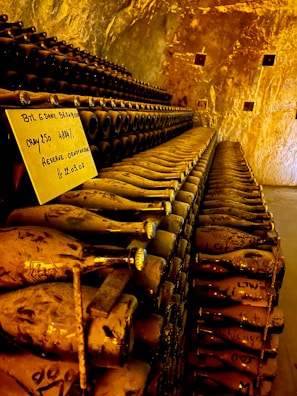A dimly lit cellar filled with rows of dusty wine bottles, all stacked horizontally on shelves. A handwritten label is placed among the bottles, indicating some handwritten notes. The golden-brown tones of the walls and lighting create a warm, aged atmosphere.