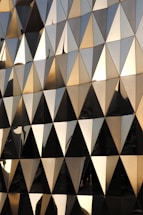 A geometric facade composed of numerous triangular panels with a reflective, metallic surface. The sunlight casts a warm glow across the panels, creating an interplay of light and shadow. The reflections hint at nearby structures and sky, adding layers of visual complexity.