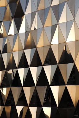A geometric facade composed of numerous triangular panels with a reflective, metallic surface. The sunlight casts a warm glow across the panels, creating an interplay of light and shadow. The reflections hint at nearby structures and sky, adding layers of visual complexity.