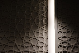 A wall featuring a geometric star pattern is highlighted by a vertical strip of light. The design has a three-dimensional effect, casting shadows on the surface and creating a sense of depth.