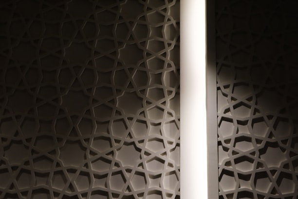 A wall featuring a geometric star pattern is highlighted by a vertical strip of light. The design has a three-dimensional effect, casting shadows on the surface and creating a sense of depth.