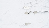 Peeling white paint on a rough-textured wall, revealing a grayish underlayer. The surface exhibits numerous cracks and flaking sections, giving a worn and aged appearance.
