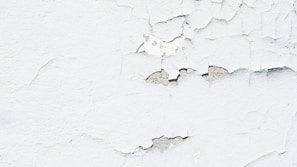 Peeling white paint on a rough-textured wall, revealing a grayish underlayer. The surface exhibits numerous cracks and flaking sections, giving a worn and aged appearance.