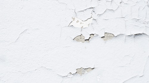 Peeling white paint on a rough-textured wall, revealing a grayish underlayer. The surface exhibits numerous cracks and flaking sections, giving a worn and aged appearance.