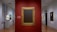 A museum gallery featuring a classic and elegant interior with smooth wooden floors and white walls. In the center is a wall painted red, showcasing a large, empty gold-framed canvas. To the left is a sculpture of a head displayed on a pedestal and a painting with a portrait on the wall. On the right, additional paintings are hung, including portraits of individuals. The gallery is well-lit with spotlights accentuating the art pieces.