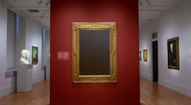 A museum gallery featuring a classic and elegant interior with smooth wooden floors and white walls. In the center is a wall painted red, showcasing a large, empty gold-framed canvas. To the left is a sculpture of a head displayed on a pedestal and a painting with a portrait on the wall. On the right, additional paintings are hung, including portraits of individuals. The gallery is well-lit with spotlights accentuating the art pieces.