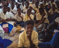 A large group of students, mostly boys, are gathered in what appears to be a classroom or assembly hall. They are sitting closely together on the floor, wearing school uniforms that include white shirts and blue trousers. Some students are holding notebooks and pens, suggesting an educational setting. The atmosphere seems studious, with many students appearing attentive or contemplative.