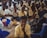 A large group of students, mostly boys, are gathered in what appears to be a classroom or assembly hall. They are sitting closely together on the floor, wearing school uniforms that include white shirts and blue trousers. Some students are holding notebooks and pens, suggesting an educational setting. The atmosphere seems studious, with many students appearing attentive or contemplative.