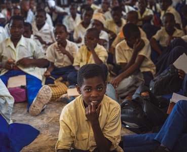 A large group of students, mostly boys, are gathered in what appears to be a classroom or assembly hall. They are sitting closely together on the floor, wearing school uniforms that include white shirts and blue trousers. Some students are holding notebooks and pens, suggesting an educational setting. The atmosphere seems studious, with many students appearing attentive or contemplative.