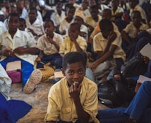 A large group of students, mostly boys, are gathered in what appears to be a classroom or assembly hall. They are sitting closely together on the floor, wearing school uniforms that include white shirts and blue trousers. Some students are holding notebooks and pens, suggesting an educational setting. The atmosphere seems studious, with many students appearing attentive or contemplative.