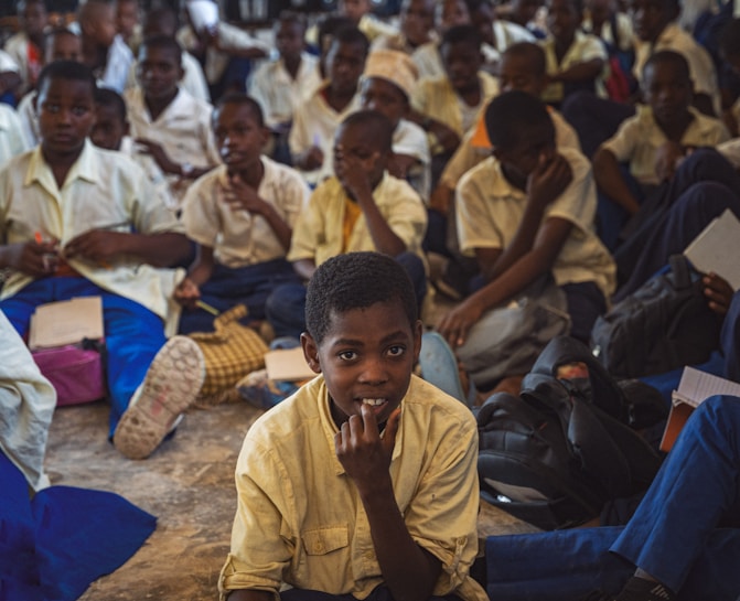 A large group of students, mostly boys, are gathered in what appears to be a classroom or assembly hall. They are sitting closely together on the floor, wearing school uniforms that include white shirts and blue trousers. Some students are holding notebooks and pens, suggesting an educational setting. The atmosphere seems studious, with many students appearing attentive or contemplative.