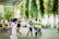 A group of young people are playing futsal on an outdoor court. Two players in bright green jerseys are hugging, possibly celebrating. Other players, wearing various team uniforms, are nearby, and there are motorcycles parked and spectators sitting in the background. The surrounding area contains green trees, providing a natural backdrop.