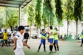 A group of young people are playing futsal on an outdoor court. Two players in bright green jerseys are hugging, possibly celebrating. Other players, wearing various team uniforms, are nearby, and there are motorcycles parked and spectators sitting in the background. The surrounding area contains green trees, providing a natural backdrop.