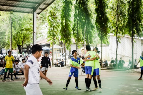 A group of young people are playing futsal on an outdoor court. Two players in bright green jerseys are hugging, possibly celebrating. Other players, wearing various team uniforms, are nearby, and there are motorcycles parked and spectators sitting in the background. The surrounding area contains green trees, providing a natural backdrop.