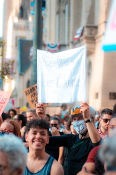 A group of people participating in a protest, with a person holding a sign that says 'Her Jam Her Choice'. The crowd includes individuals of different ages and genders, and some are wearing masks. The background shows a building with architectural details and flags.