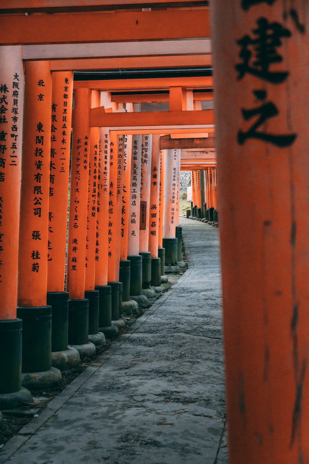 a corridor with orange and black pillars with Fushimi Inari-taisha in the background