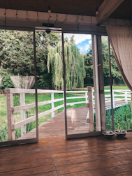 A spacious indoor area features floor-to-ceiling sliding glass doors opening to a view of a lush garden. A wooden deck leads to a white wooden fence that borders a grassy area with a willow tree. Two metal pet bowls are placed on the wooden floor inside.