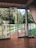 A spacious indoor area features floor-to-ceiling sliding glass doors opening to a view of a lush garden. A wooden deck leads to a white wooden fence that borders a grassy area with a willow tree. Two metal pet bowls are placed on the wooden floor inside.