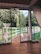 A spacious indoor area features floor-to-ceiling sliding glass doors opening to a view of a lush garden. A wooden deck leads to a white wooden fence that borders a grassy area with a willow tree. Two metal pet bowls are placed on the wooden floor inside.