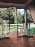 A spacious indoor area features floor-to-ceiling sliding glass doors opening to a view of a lush garden. A wooden deck leads to a white wooden fence that borders a grassy area with a willow tree. Two metal pet bowls are placed on the wooden floor inside.