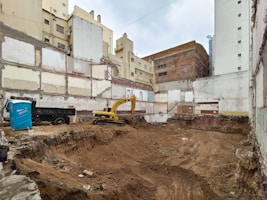 A construction site is set within a cityscape, with excavation activities in progress. Exposed, unfinished buildings serve as the backdrop, and walls display signs of wear and decay. A yellow excavator is actively engaged in digging, and a blue portable toilet is situated nearby. The surroundings appear urban, with several tall buildings partially visible.