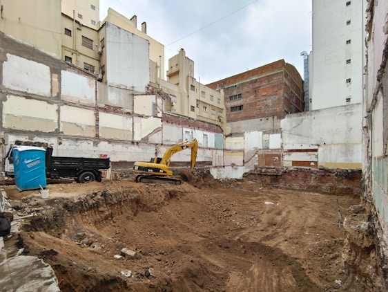A construction site is set within a cityscape, with excavation activities in progress. Exposed, unfinished buildings serve as the backdrop, and walls display signs of wear and decay. A yellow excavator is actively engaged in digging, and a blue portable toilet is situated nearby. The surroundings appear urban, with several tall buildings partially visible.