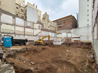A construction site is set within a cityscape, with excavation activities in progress. Exposed, unfinished buildings serve as the backdrop, and walls display signs of wear and decay. A yellow excavator is actively engaged in digging, and a blue portable toilet is situated nearby. The surroundings appear urban, with several tall buildings partially visible.