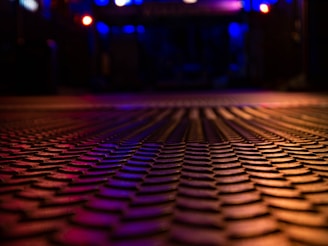 A textured surface with a repetitive pattern fills the foreground, emphasizing a sense of depth. The scene is bathed in low ambient lighting, with blue and red tones lending a moody, atmospheric feel. The background is softly blurred but hints at an urban or industrial setting with colorful lights.