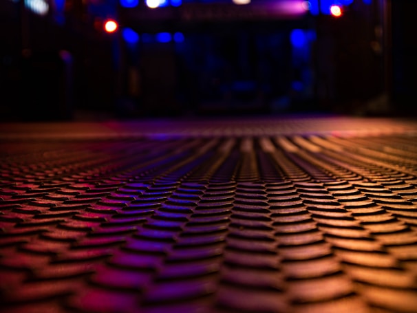 A textured surface with a repetitive pattern fills the foreground, emphasizing a sense of depth. The scene is bathed in low ambient lighting, with blue and red tones lending a moody, atmospheric feel. The background is softly blurred but hints at an urban or industrial setting with colorful lights.